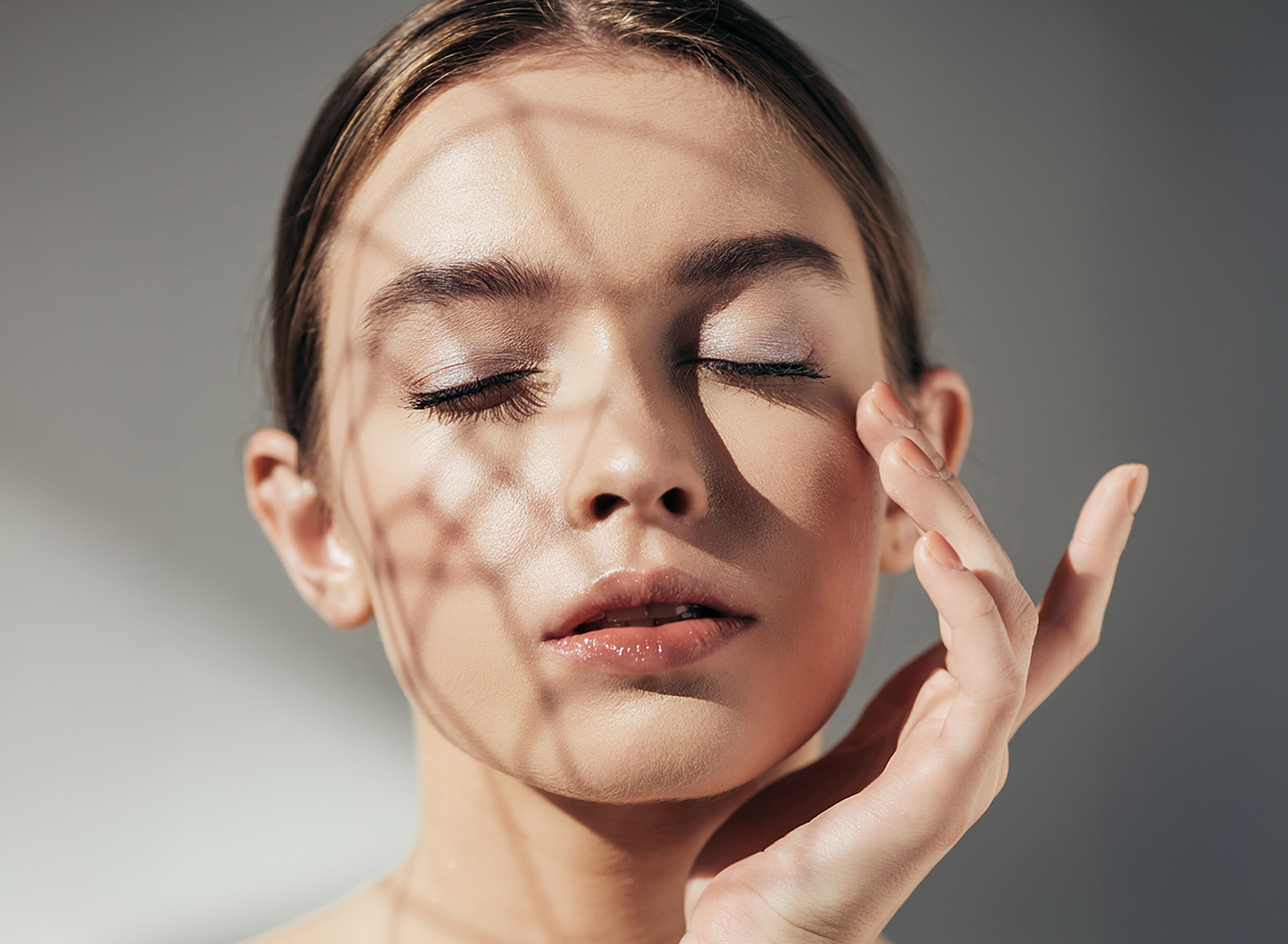 Porträt einer Frau mit geschlossenen Augen, sanftem Licht- und Schattenspiel im Gesicht, die mit den Fingern ihr Augenlid berührt – Ausdruck von ruhiger, gepflegter Haut und Augenpartie.