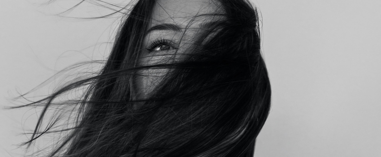 Schwarz-Weiß-Aufnahme einer Frau, deren Gesicht teilweise von ihren im Wind wehenden Haaren verdeckt wird.