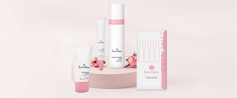 Unterschiedliche Pflegeprodukte auf einem rosafarbenem Podest mit dekorativen Blüten.