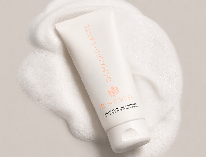 Tube der Jean D'Arcel DÉMAQUILLANTE Anti-Aging Cleansing Mousse auf weichem Reinigungsschaum vor hellem Hintergrund.